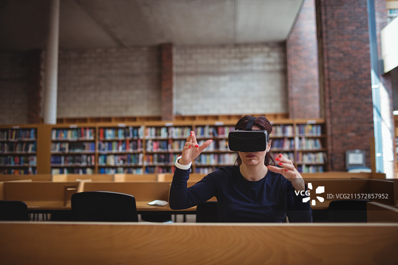 女人在图书馆的阅读室里佩戴虚拟现实头盔，双手举起，坐在木桌前。图片素材