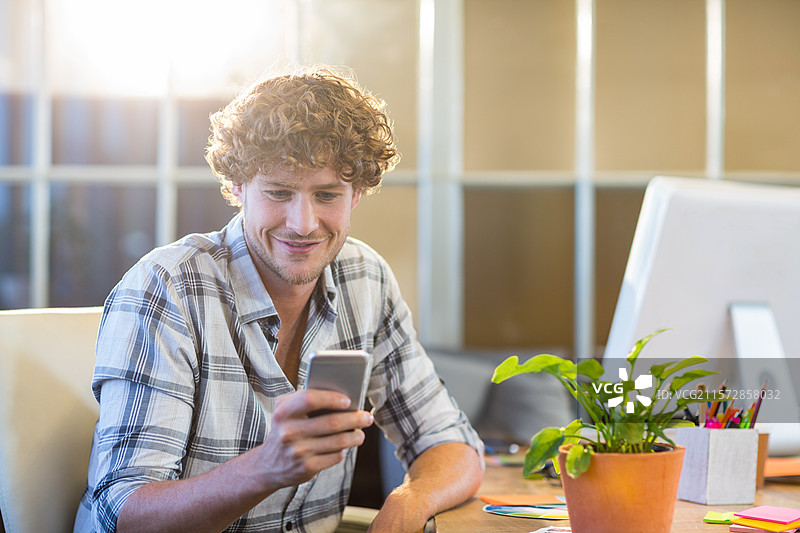男子坐在木桌前，手持智能手机，桌面上有显示器和盆栽植物。图片素材