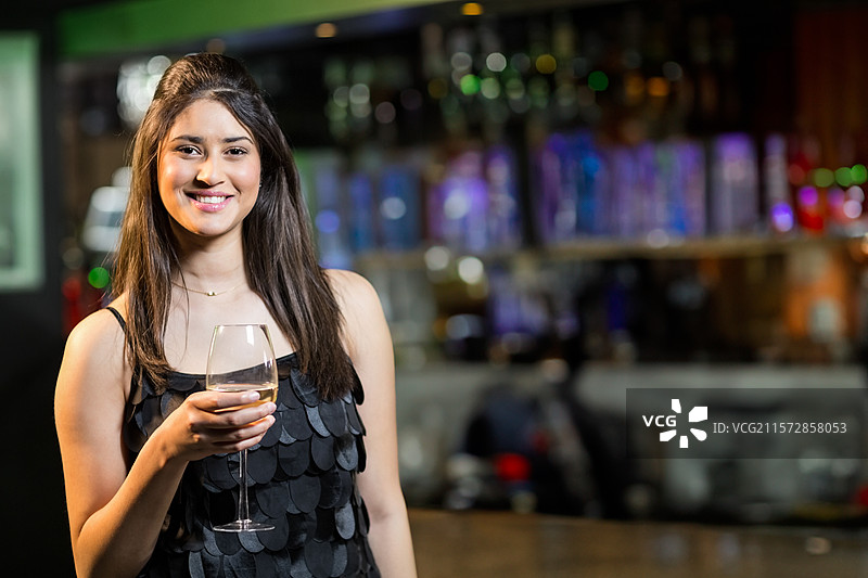 穿着黑色连衣裙的女人在酒吧里举着酒杯，周围是点亮的酒瓶和柔和的灯光，留有空白空间。图片素材