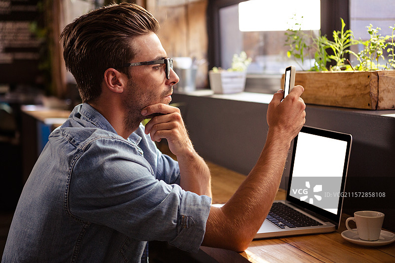 智能手机静静地放在大窗户旁的木质台面上，旁边有笔记本电脑、咖啡杯和盆栽植物。图片素材