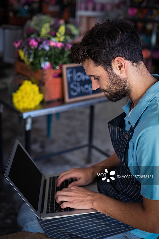 留着胡子的花店老板在花店的工作区用笔记本电脑打字，周围是装满五彩缤纷花朵的木箱。图片素材