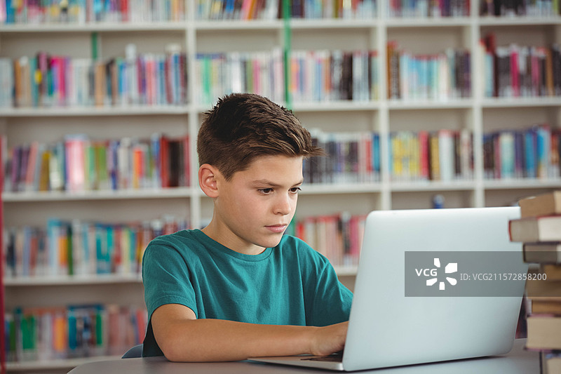 男孩在学校图书馆的阅读室里，桌子上用银色笔记本电脑学习，周围是白色书架。图片素材