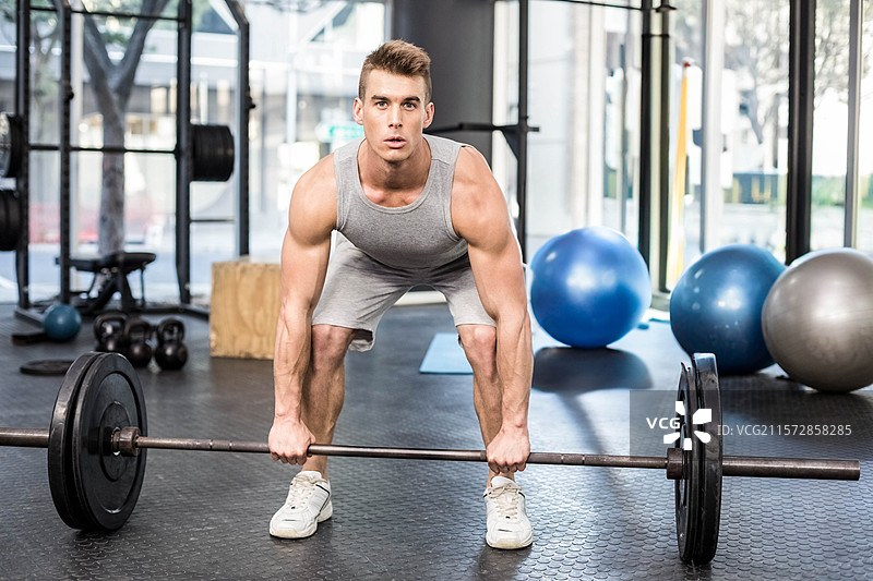 肌肉壮男在垫子上进行硬拉，握住杠铃，旁边是窗边的壶铃，窗外可见树木。图片素材