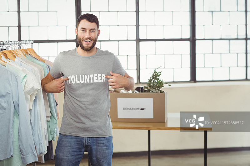 微笑着指着志愿者T恤的男性志愿者，站在慈善房间的捐款箱旁。图片素材