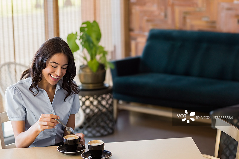 二十多岁的女性在咖啡馆休息区的桌子上搅拌咖啡，桌上有黑色咖啡杯和盆栽植物。图片素材
