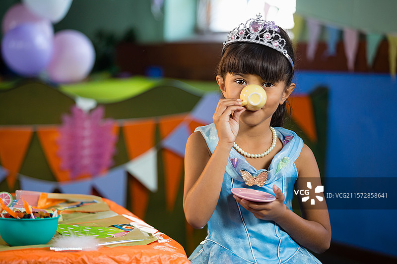 女孩在派对桌旁用粉色茶托喝黄色玩具茶杯里的饮料，周围有柔和色调的气球。图片素材