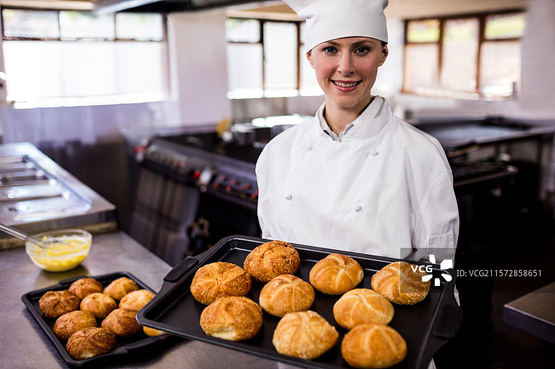 穿着厨师外套和厨师帽的女厨师，在餐厅厨房里手持装有金黄色面包卷的烤盘图片素材