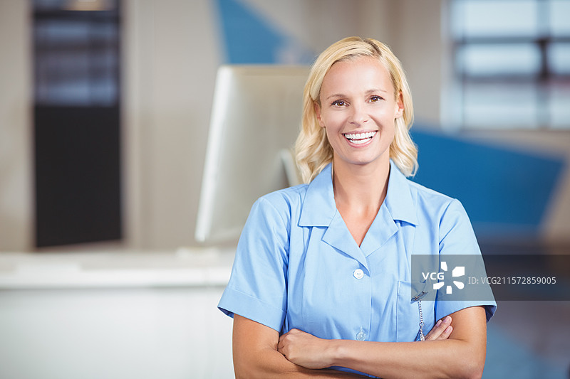 微笑的女性护士穿着蓝色护士服，双臂交叉站在诊所接待处，旁边有电脑显示器。图片素材