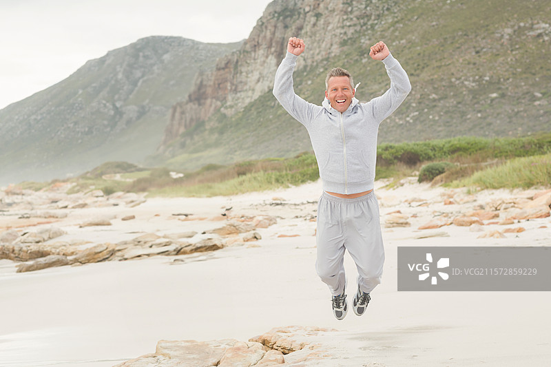男子在沙滩上空中跳跃，穿着浅灰色拉链连帽衫、宽松运动裤和运动鞋。图片素材