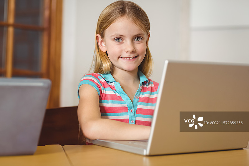 小女孩坐在木质书桌前，微笑着直视镜头，使用银色笔记本电脑。图片素材
