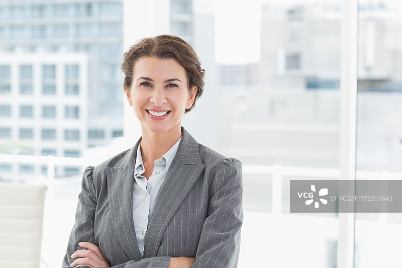穿着条纹西装的女商人，双臂交叉在办公椅旁，窗外可见城市天际线。图片素材