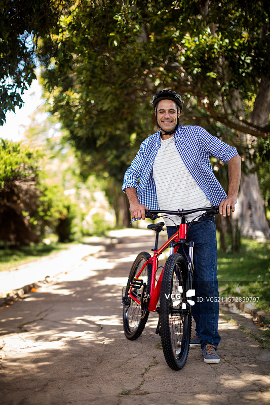 男子站在阳光明媚的树荫小道上，手持红色山地自行车，佩戴骑行头盔。图片素材