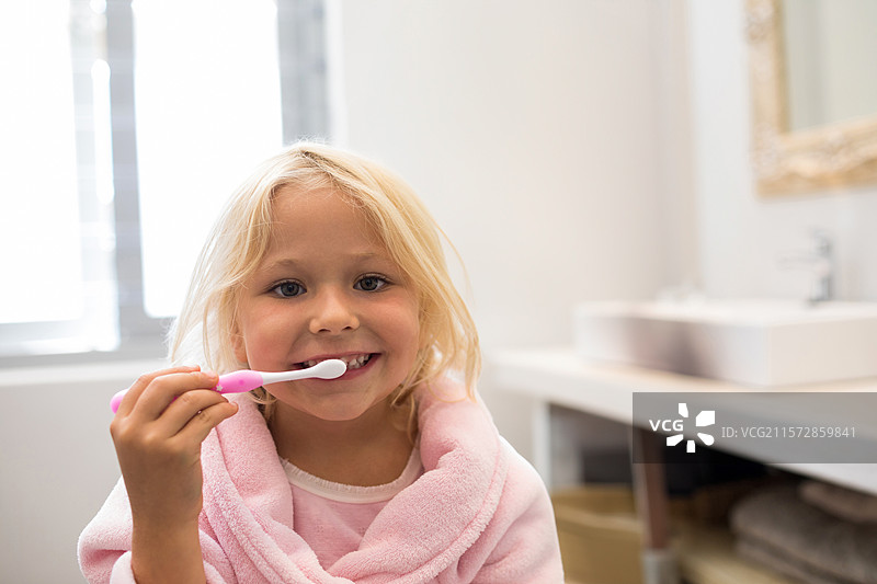 穿着粉色浴袍的女孩在浴室洗手池前刷牙，手里拿着粉色牙刷，旁边有一面金色镜子。图片素材