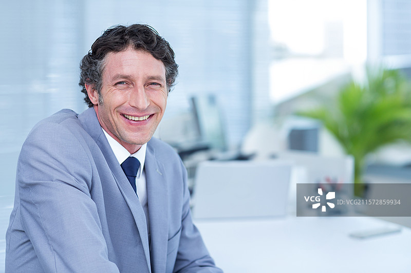 商人在办公室的白色桌子前坐着微笑，桌上有电脑显示器和绿色植物。图片素材