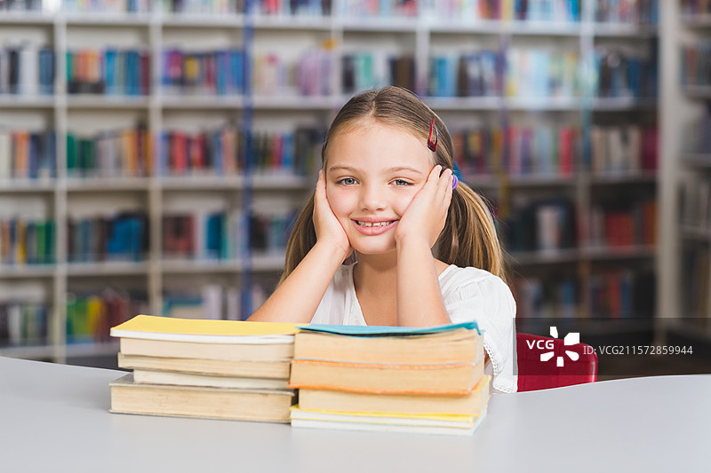 女学生在图书馆的大桌子上微笑着阅读堆叠的精装书籍图片素材