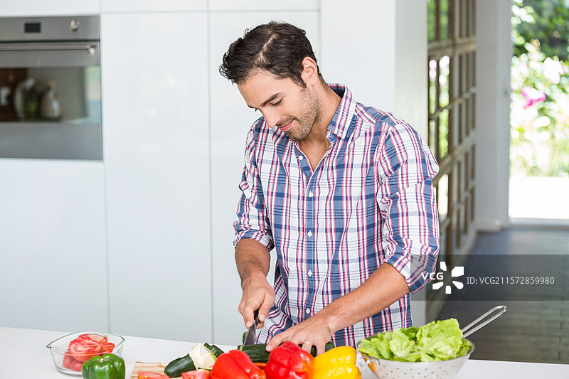 男子在家厨房的台面上用刀切西葫芦，旁边有甜椒和番茄，留有空白空间。图片素材