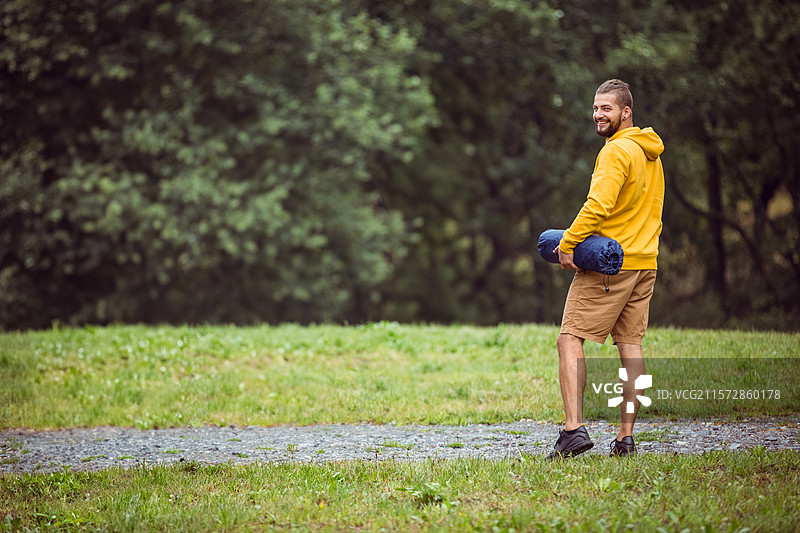 穿着芥末色连帽衫的男子站在森林边缘的空地上，手里拿着卷起的露营垫，留有空白空间。图片素材