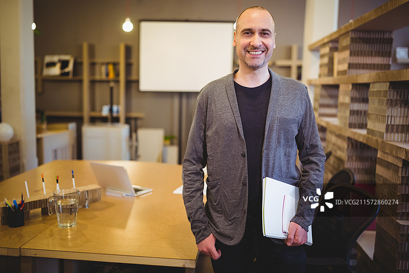 在现代办公室会议区站着微笑的男子，手里拿着文件和笔记本，旁边有一台笔记本电脑。图片素材