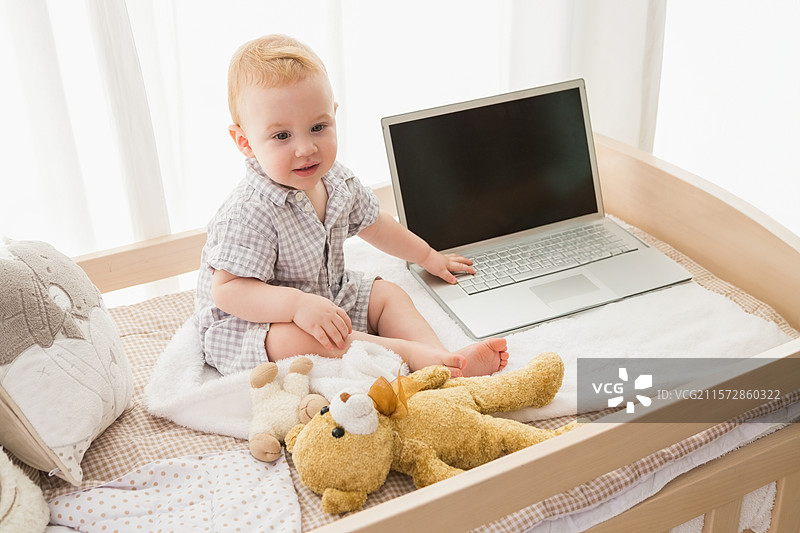 坐在木制幼儿床上的婴儿男孩直立着，手里抱着泰迪熊，旁边有一台打开的笔记本电脑和一个毛绒玩具。图片素材