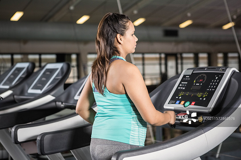 穿着背心和紧身裤的女性站在跑步机上，握住扶手查看控制台，正在健身房锻炼。图片素材