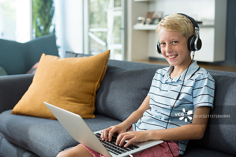 男孩穿着条纹 polo 衫和耳机，坐在家里的沙发上，正在用笔记本电脑打字，周围有靠垫。图片素材