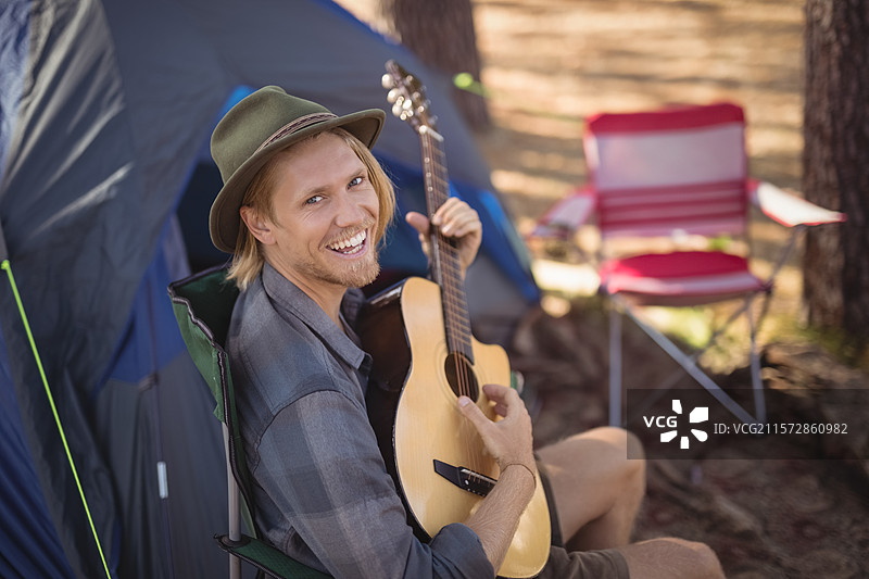 男人坐在绿色椅子上弹奏木吉他，营地里有帐篷和红白相间的条纹椅子。图片素材