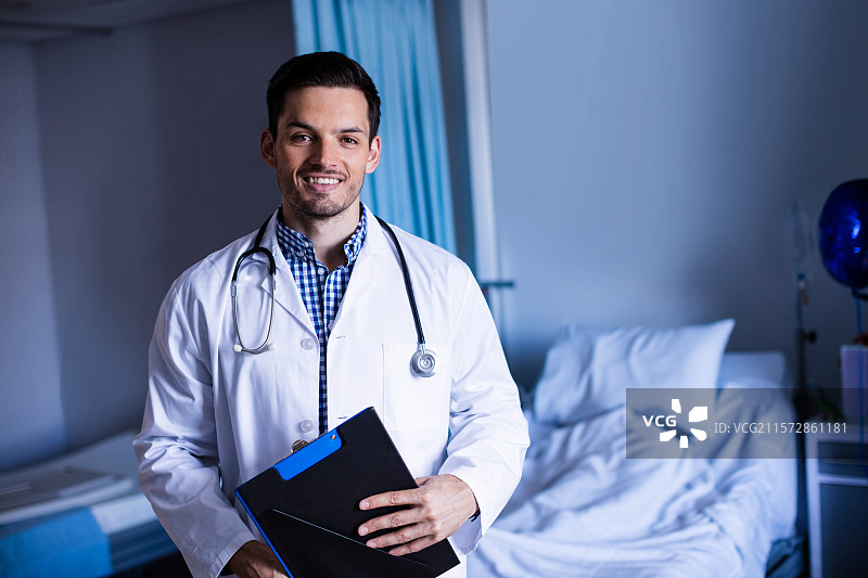 在医院病房里，一位亚洲男性医生手持夹板，佩戴听诊器。图片素材