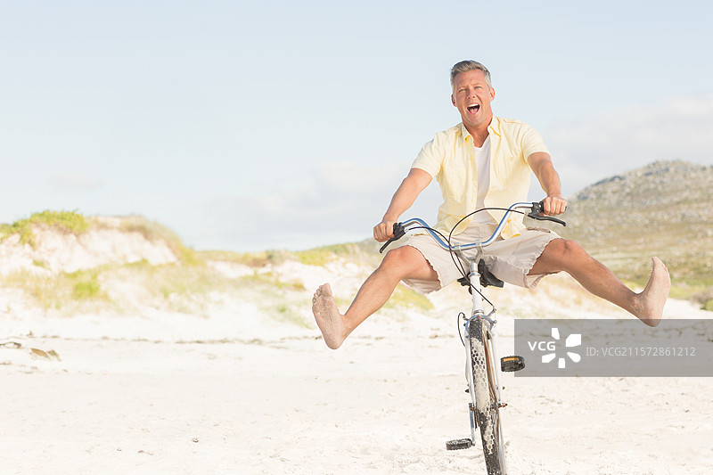 中年男子骑着沙滩自行车沿着沙丘骑行，双腿抬起，开心地大笑。图片素材