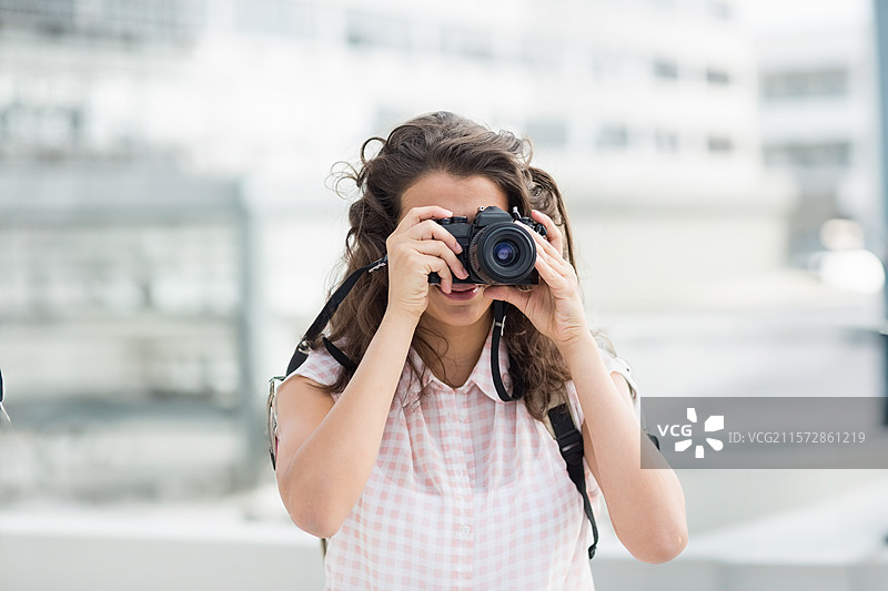 女性摄影师在屋顶露台上举起单反相机，金属栏杆和模糊的建筑外立面作为背景。图片素材