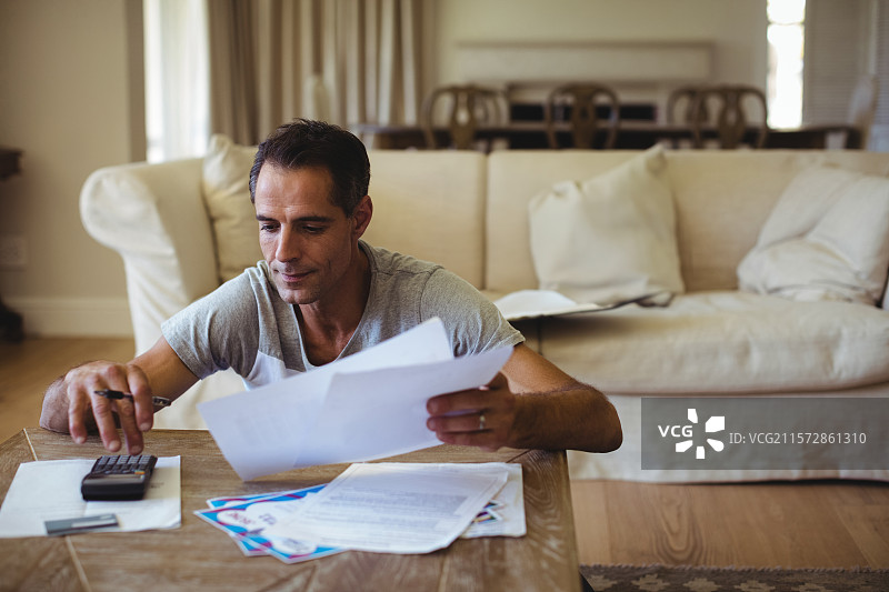 男人坐在客厅的咖啡桌旁，用计算器计算，桌上有纸张、信封和钢笔。图片素材