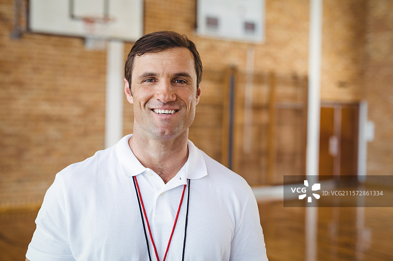 男教练站在学校体育馆里微笑，佩戴红黑色挂绳和哨子。图片素材