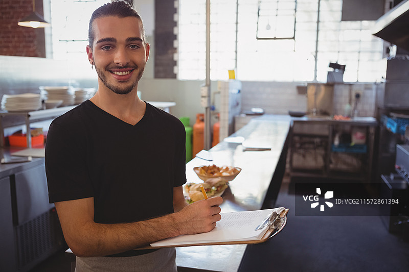 男子在餐厅厨房里手持笔和夹子，检查订单，周围有碗和盘子。图片素材