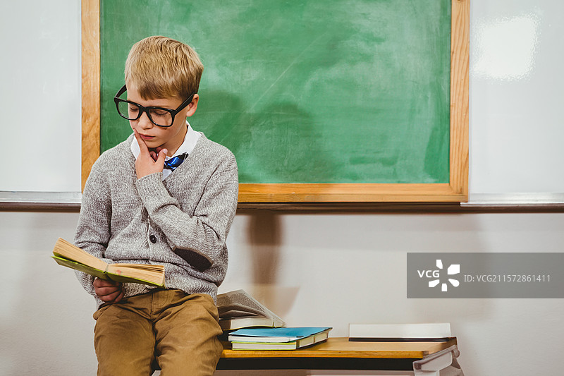 戴眼镜的学龄男孩在黑板前阅读书籍，旁边是一堆书。图片素材