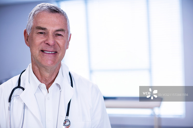 穿着白大褂和听诊器的老年男性医生站在检查室的桌子旁，留有空白空间图片素材
