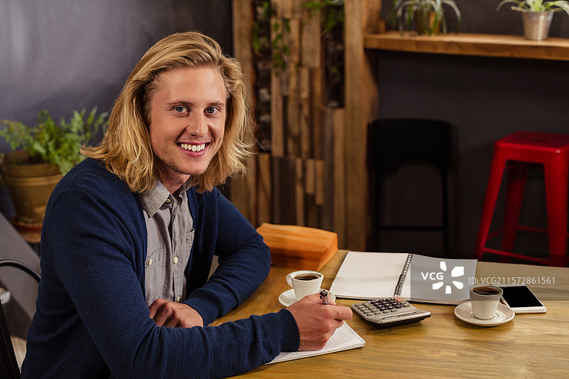 男子在咖啡馆的桌子上用笔在笔记本上写字，同时使用计算器，桌上还有咖啡杯和智能手机。图片素材