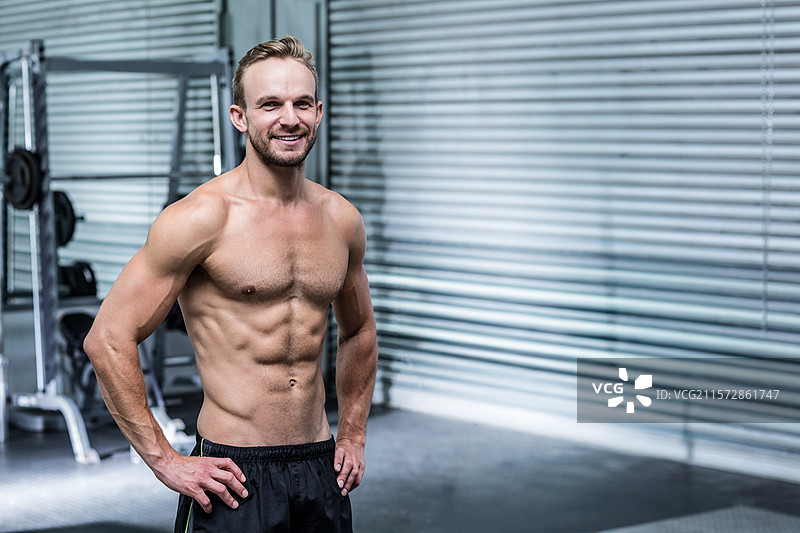 在健身房里，微笑着面对镜头的健壮男士，身旁有举重器械和金属卷帘门，留有空白空间。图片素材
