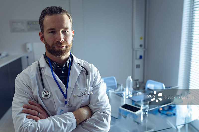 男医生穿着白大褂和听诊器，双臂交叉站在诊所办公室旁的笔记本电脑前。图片素材