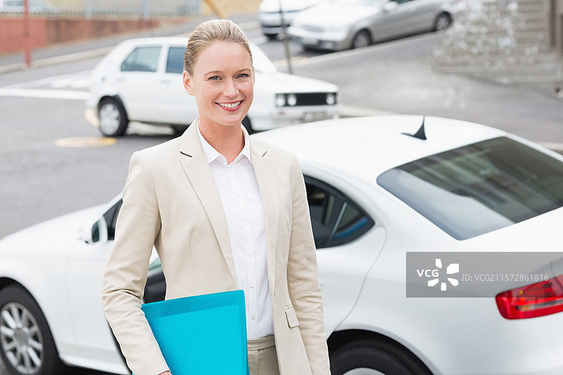 女商人在城市街道旁站着，微笑着手持蓝色文件夹，旁边是一辆白色轿车和一辆掀背车。图片素材