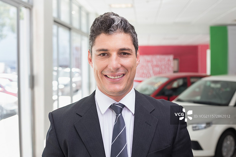 穿着西装和条纹领带的中年男子站在玻璃窗旁的汽车展厅内。图片素材