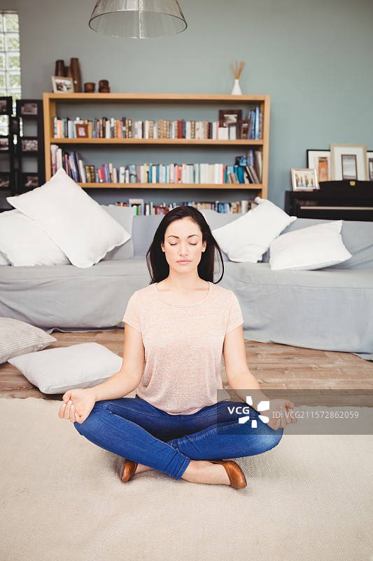 女人盘腿坐在客厅的米色地毯上冥想，身旁有书架。图片素材