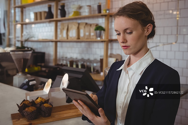 穿着西装的女性在咖啡馆的柜台后使用平板电脑，靠近收银机和咖啡研磨机，留有空白空间。图片素材