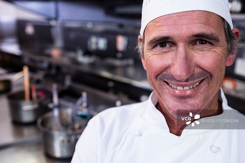 微笑的厨师穿着白色外套，站在钢制台面旁，周围有锅、平底锅和餐具，悬挂灯光下。图片素材