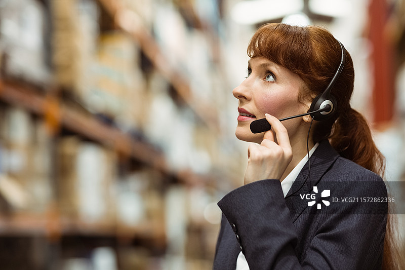 穿着西装的女商人在仓库中佩戴耳机检查库存，旁边是整齐的箱子，留有空白空间。图片素材