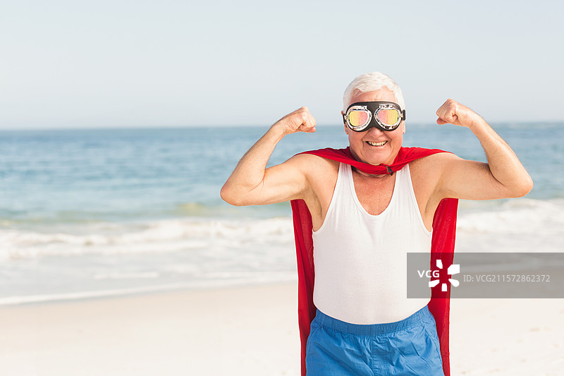 一位老年男子在沙滩上展示手臂，身穿红色斗篷，佩戴反光护目镜，背景是海浪。图片素材