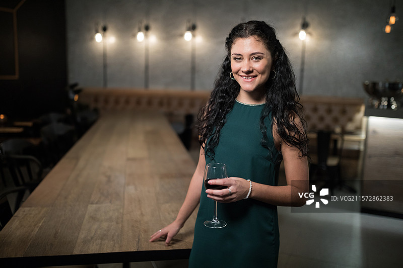 穿着绿色连衣裙的女人靠在酒吧的桌子上，手中拿着一杯红酒，留有空白空间。图片素材