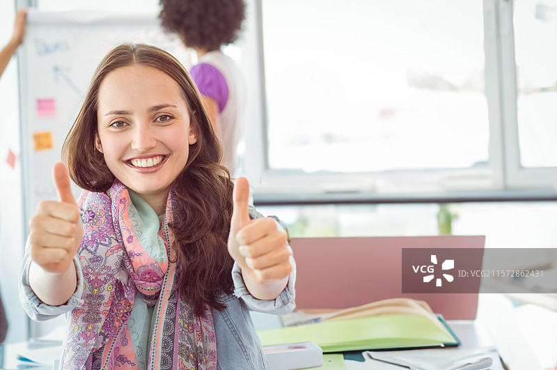 微笑的成年女性工作人员坐在桌子旁，给出竖起大拇指的手势，桌上有笔记本电脑和白板笔记。图片素材