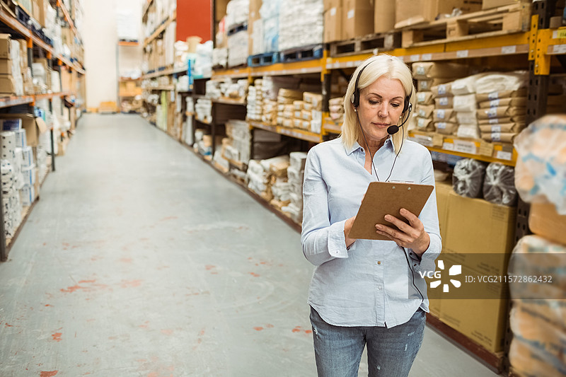 女性站在仓库过道中，手持夹板扫描库存，佩戴耳机，留有空白空间。图片素材