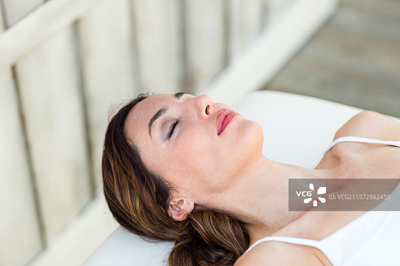 女人穿着吊带衫，躺在水疗甲板上舒适的治疗台上，靠在靠垫上，旁边是栏杆。图片素材
