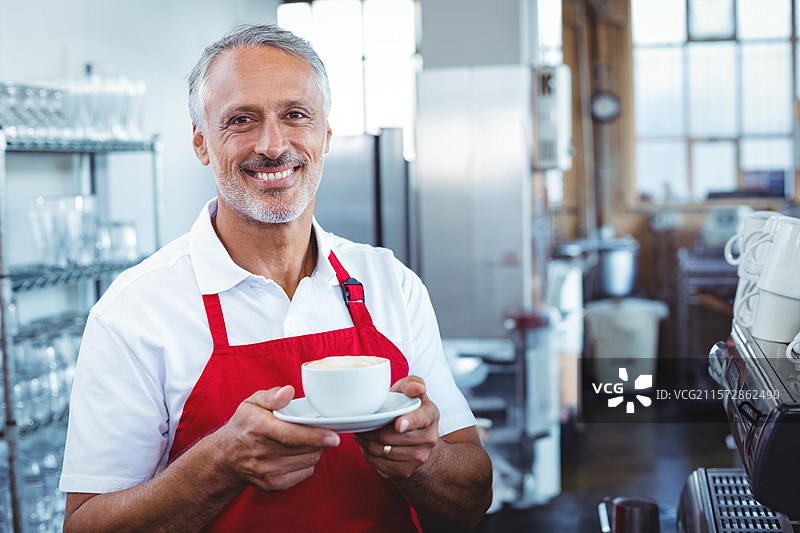 在咖啡馆里，穿着围裙的老年男咖啡师在浓缩咖啡机后面，手里端着一个杯子和碟子。图片素材