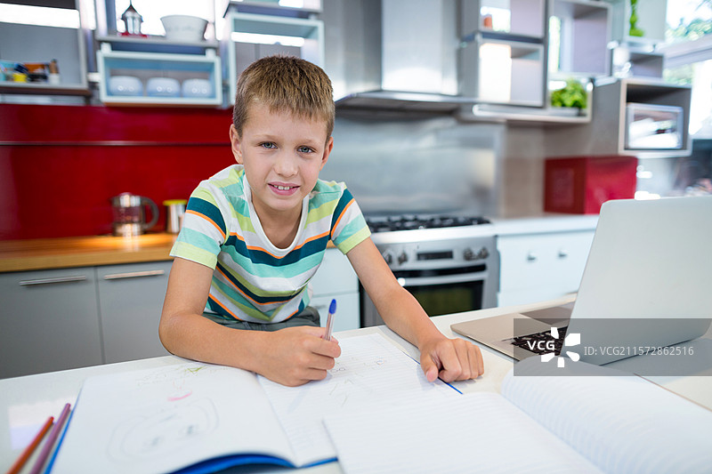 在厨房台面上用蓝色钢笔写笔记本的学龄男孩，旁边有笔记本电脑和彩色铅笔。图片素材